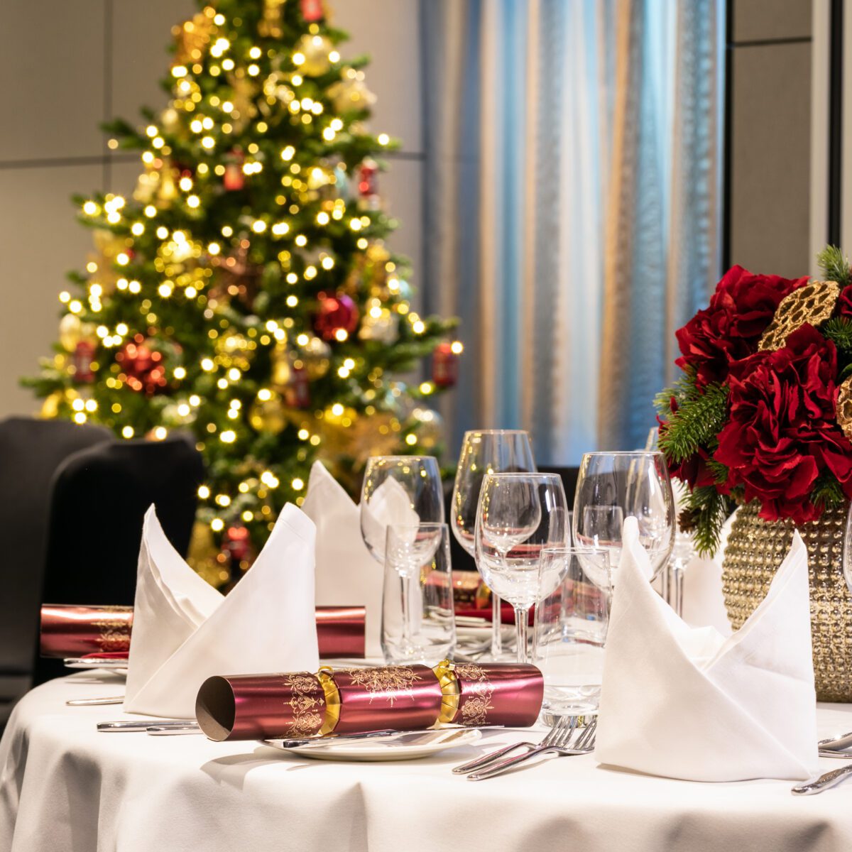 A table with glasses, crackers, and flowers in front of of a lit Christmas tree.